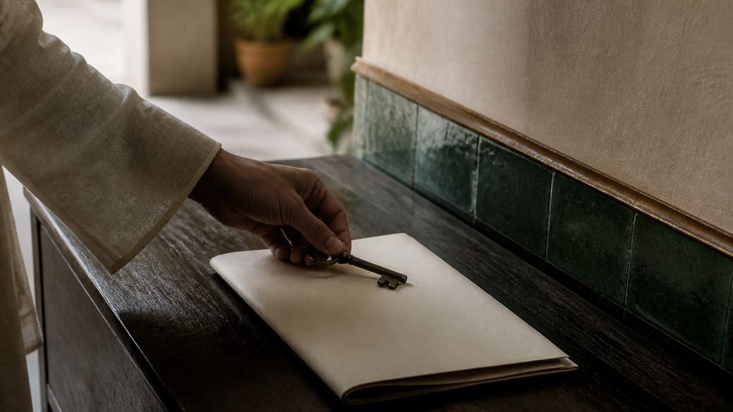 Main anonyme déposant une clé sur un folio ivoire sans texte, entre bois sombre, zellige vert et laiton patiné.
