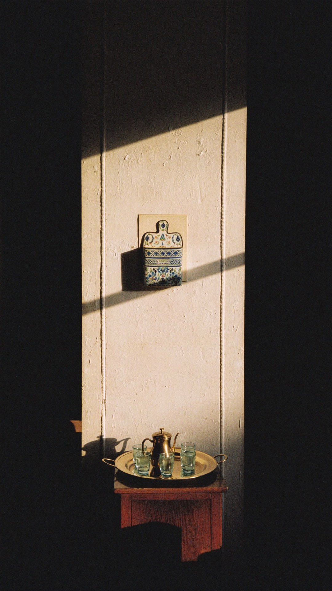 Détail : un plateau de thé posé sur un tabouret, traversé par une lame de lumière sur un mur clair.